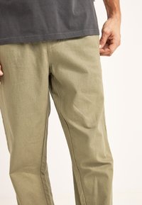 Los pantalones caqui presentan una superficie lisa y texturizada con un diseño de pierna recta y costuras laterales sutiles. Combinados con una camisa gris oscuro, visible en la parte superior.