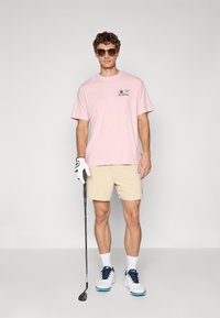 Lys rosa bomulls t-skjorte med et lappedesign, beige shorts, hvite golf sko med blå detaljer, og solbriller. Golfkølle i hånden.