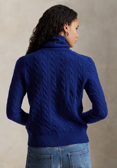 Blå kabelstrikket genser med høy hals og ribbestrikket kant. Har strukturert mønster og lange ermer, brukt over denim.