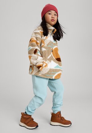 Beige Fleece-Pullover mit abstrakten Mustern in Orange, Braun und Türkis; kombiniert mit hellblauen Hosen und braunen Knöchelstiefeln.