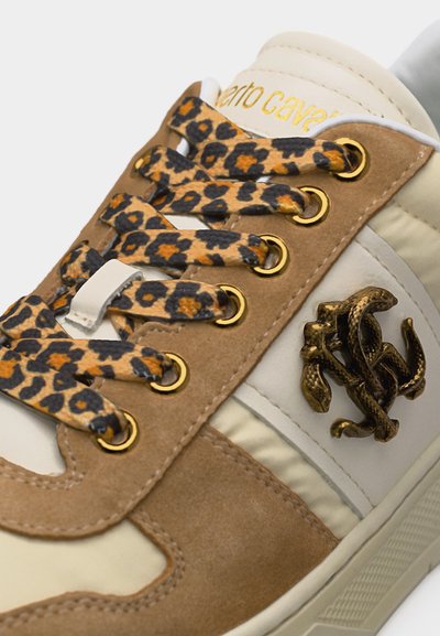 Κοντινό πλάνο μπεζ και λευκού αθλητικού παπουτσιού με καφέ σουέντ λεπτομέρειες, κορδόνια με λεοπάρ σχέδιο, χρυσά μεταλλικά τρύπες για κορδόνια και μεταλλικό λογότυπο σε σχήμα φιδιού στο πλάι.