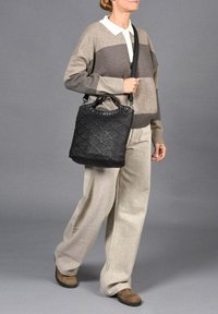 Zwarte geweven handtas met een gestructureerd oppervlak, voorzien van ronde handvatten en een afneembare schouderriem, gedragen met een grijs-bruin gestreept sweater en beige broek.