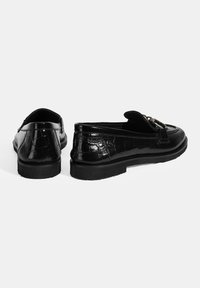Zwarte gepatineerde leren loafers met een slank ontwerp, ronde neus en decoratieve hardwaredetail. Ruime gladde textuur en lage zool.