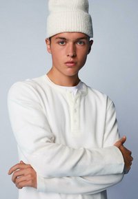 Weißes, geripptes Langarm-Henley-Shirt mit drei Knöpfen am Kragen, kombiniert mit einer weißen Strickmütze, vor einem hellblauen Hintergrund.