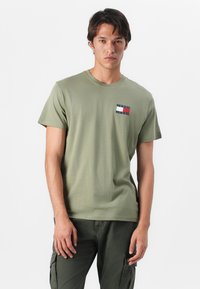 Olive green cotton t-shirt with a crew neckline and short sleeves, featuring a logo patch with red, white, and blue colors on the chest.