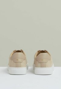 Beige semišové tenisky s bílými gumovými podrážkami, mají nízký střih a diskrétní zlatou značku na zadní straně. Celkově hladká textura.