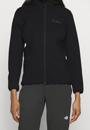 Personne portant une veste zippée noire Jack Wolfskin et un pantalon noir The North Face, debout, les bras détendus le long du corps.