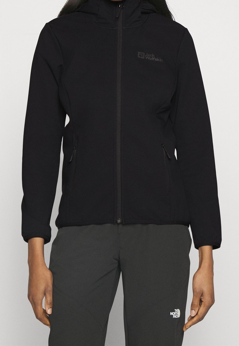 Personne portant une veste zippée noire Jack Wolfskin et un pantalon noir The North Face, debout, les bras détendus le long du corps.