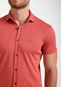 Trumpas rankovės polo marškinėliai šviesiai koralinės spalvos, pagaminti iš lygaus, elastingo audinio. Savybės: mygtukiniai atvartai ir mažas logotipas ant krūtinės.