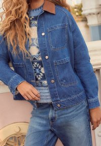 Kurtka dżinsowa z brązowym kołnierzem z kordury, z dwiema kieszeniami na piersiach i zapięciem na guziki, noszona na wzorzystym swetrze i niebieskich dżinsach.