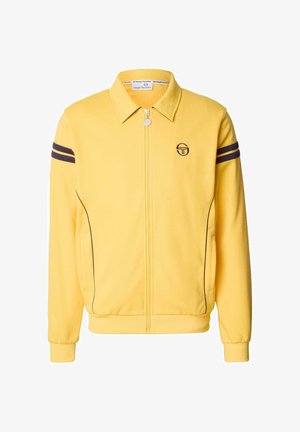 Chaqueta amarilla con cremallera y cuello, con rayas marinas en las mangas y un logo bordado en el pecho. Textura de tela suave.