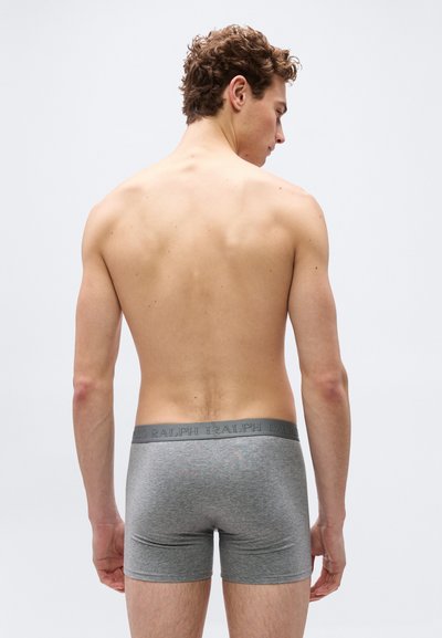 Άνδρας με σγουρά μαλλιά που κοιτάζει προς τα πίσω, φοράει γκρι εσώρουχα boxer με λωρίδα μέσης με λογότυπο, σε ένα απλό φόντο.