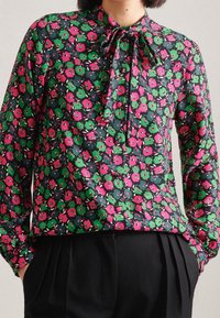 Blusa a maniche lunghe su sfondo blu navy con un motivo floreale di fiori rosa e verdi, con colletto a fiocco e una texture di tessuto liscia.