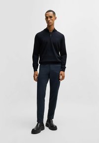 Maglione zip-up nero con una texture liscia, abbinato a pantaloni attillati blu scuro e stivaletti neri con suola spessa. Design minimalista.