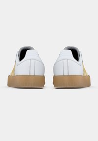 Białe tenisówki z żółtymi akcentami, teksturowaną gumową podeszwą i prążkowanym wykończeniem z tyłu. Gładki materiał wierzchni o strukturalnym wzorze.