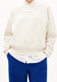 Felpa color crema con colletto alto e un piccolo logo "ARMEDANGELS" al centro. Indossata sopra una camicia bianca e pantaloni blu.