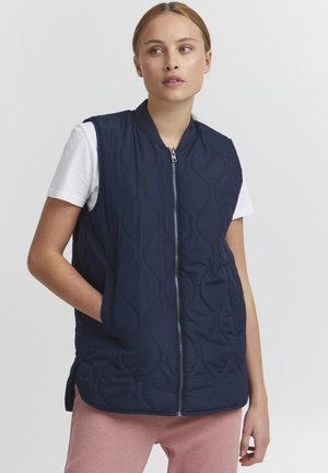 Gilet matelassé bleu marine avec fermeture éclair à l'avant, col mandarin et poches latérales, porté sur un t-shirt blanc et un pantalon rose. Texture de tissu lisse.