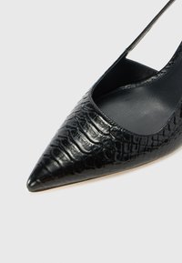 Scarpa con tacco alto in pelle verniciata nera, punta affusolata e finitura in pelle di coccodrillo, caratterizzata da un taglio basso e un design con cinturino sottile alla caviglia.