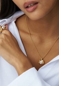 Κολιέ με χρυσό μενταγιόν και στρογγυλή πέρλα στο κέντρο, με λείο, στρογγυλό σχέδιο. Φοριέται πάνω από ένα απλό λευκό πουκάμισο.