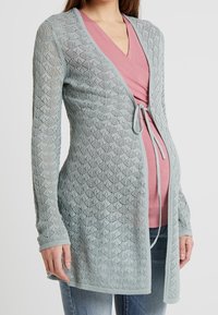Cardigan en tricot bleu clair avec un motif en diamants, doté d'une fermeture à nouer à la taille et de manches longues, porté sur un haut rose et un jean.