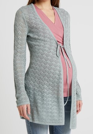 Lichtblauwe gebreide cardigan met een diamantpatroon, voorzien van een striksluiting in de taille en lange mouwen, gedragen over een roze top en jeans.