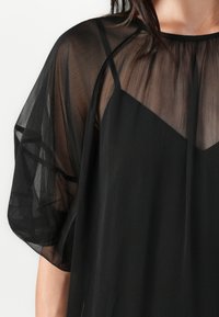 Černá průsvitná halenka s měkkou, splývavou látkou. Má uvolněný střih, široké rukávy a vrstvený design s matnou spodní vrstvou.