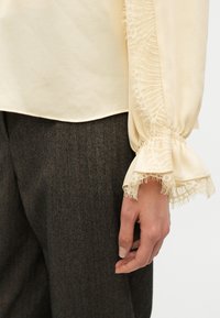 Manche de blouse crème avec bordure en dentelle et poignet froncé à côté d’un pantalon en laine gris foncé à motif chevrons, montrant une position de main détendue.