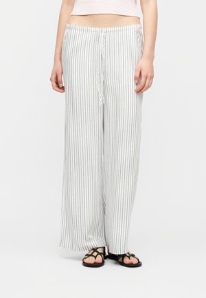 Osoba oblečená v voľných bielych nohaviciach s vertikálnymi sivými pruhmi a čiernych sandáloch s remienkami stojaca pred jednoduchým pozadím.