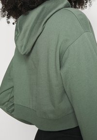 Persona con capelli ricci che indossa una felpa corta color verde oliva con maniche lunghe e orlo a coste, mostrata di spalle.