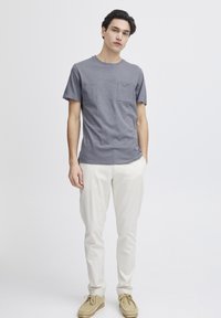 Tricou gri cu mânecă scurtă, dotat cu un buzunar pe piept, purtat cu pantaloni bej deschis și pantofi pe nuanță de tan, pe un fundal alb simplu.