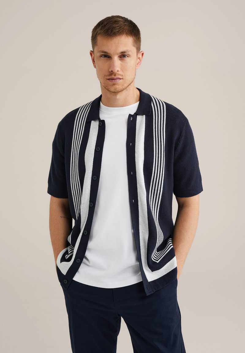 Cárdigan de manga corta en color navy con un diseño de rayas blancas que corre verticalmente por la parte delantera, cuenta con cierre de botones y una textura suave.