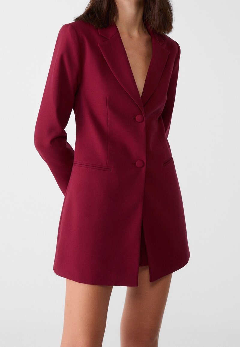 Femme portant une robe blazer rouge profond ajustée avec manches longues et deux boutons, debout devant un fond blanc uni.