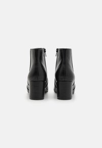 Botas de cuero negro hasta el tobillo con un tacón grueso, que cuentan con dos cremalleras laterales y una textura suave. El diseño es elegante y moderno.