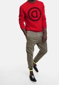 Sudadera roja con un logo circular negro, combinada con pantalones cargo verde oliva con bolsillos y dobladillo con puño. Zapatillas negras con suelas blancas.