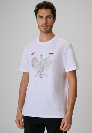 Mężczyzna w białej koszulce z srebrnym geometrycznym orłem, flagą Polski i czerwonym logo 4F na jednolitym szarym tle.