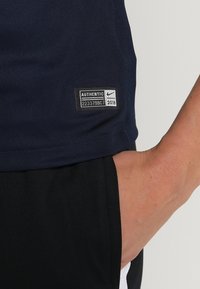 Primer plano de la mano de una persona descansando en la cadera, vistiendo una camisa azul marino con una etiqueta negra de "Authentic Nike 2018" y pantalones negros.