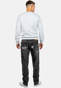 Jeans in denim nero con cuciture bianche intricate e decorazioni sulle tasche. Modello che indossa una felpa grigia chiara, visto da dietro.