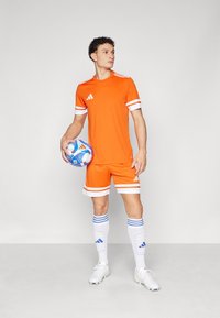 Camiseta y pantalones cortos deportivos naranjas con rayas blancas, combinados con calcetines blancos a rayas azules y botas de fútbol blancas, sosteniendo un balón de fútbol azul.