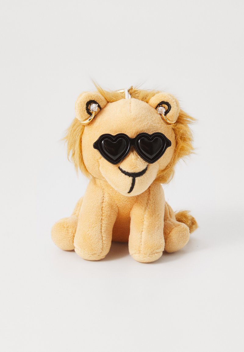 Mehka igrača lev s rumeno-rjavimi odtenki, ki nosi črne sončne očale v obliki srca ter ima puhasto grivo in majhne ušesne akcentne detajle.