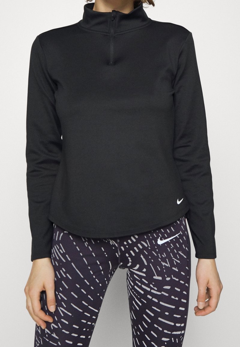 Femme portant un haut noir à manches longues avec fermeture éclair au quart et un legging noir avec des motifs abstraits blancs et des logos Nike.