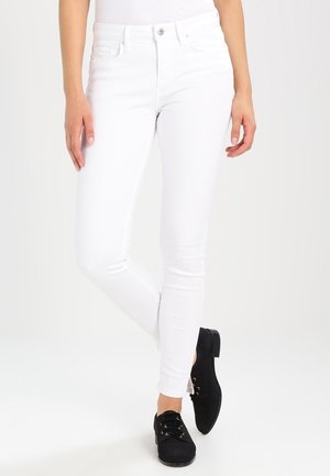 Vrouw draagt slanke witte jeans met hoge taille en voorzakken, en zwarte veterschoenen, staand met één been voor het andere gekruist.