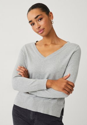 Light gray, lightweight sweater with a V-neckline, ribbed cuffs, and side slits. Soft texture, fitted design, styled with dark jeans.