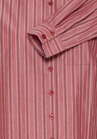 Chemise rouge à manches longues avec des rayures blanches verticales, des boutons rouges sur le devant et aux poignets, et une poche poitrine côté droit.