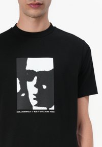 Juodas medvilninis marškinėlis su grafiniu atvaizdu, vaizduojančiu veidą balta ir pilka spalvomis, o apačioje užrašas "KARL LAGERFELD 21 RUE ST-GUILLAUME PARIS".