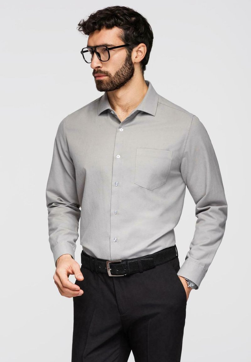 Homme avec barbe et lunettes portant une chemise gris clair boutonnée et un pantalon noir avec une ceinture noire, posant avec une main dans la poche.