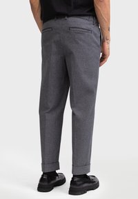 Grijze, gestructureerde broek met een losse pasvorm, voorzien van een omslagen zoom en twee achterzakken. Gecombineerd met zwarte schoenen, gezien van achteren.