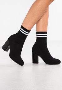 Bottines en tricot noires à bout pointu et talon bloc, avec un design de chaussette à côtes pour la cheville ornée de deux rayures blanches en haut.
