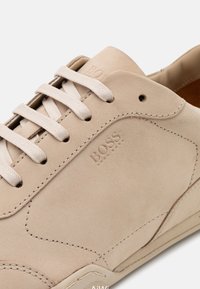 Beige semišové tenisky s plochými béžovými tkaničkami, texturovaným svrškem, tónovaným šitím a zlatou embosovanou značkou "BOSS" na boku. Viditelná gumová podrážka.