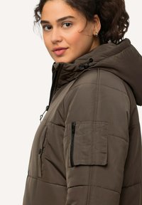 Chaqueta de plumas marrón con capucha, cierre zipper en la parte frontal y bolsillos laterales. Tejido texturizado con patrón acolchado y cordones ajustables.