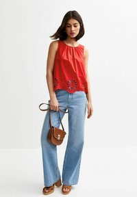 Rotes ärmelloses Oberteil mit gerafftem Ausschnitt und floralen Cut-out-Details am Saum, kombiniert mit hellblauen weit geschnittenen Jeans und einer kleinen braunen Tasche.